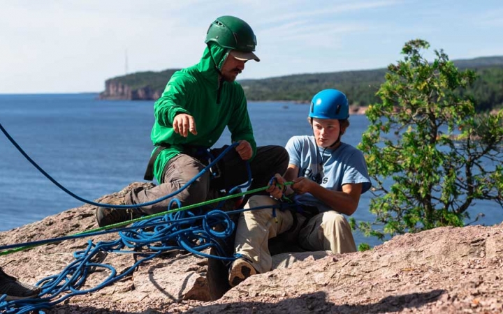 Two people wearing safety gear are secured by ropes near the edge of a cliff, above a blue body of water. One of them appears to be an instructor, giving direction to a student.